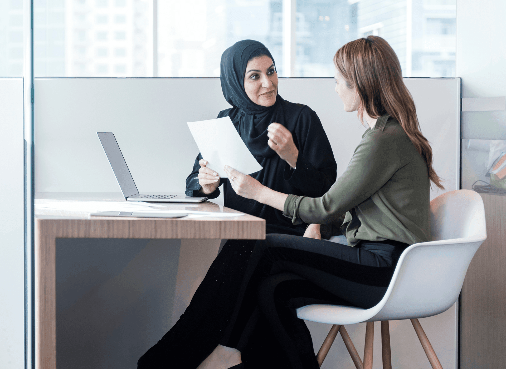 Two women discuss a document in an office setting, sitting at a desk with an open laptop.