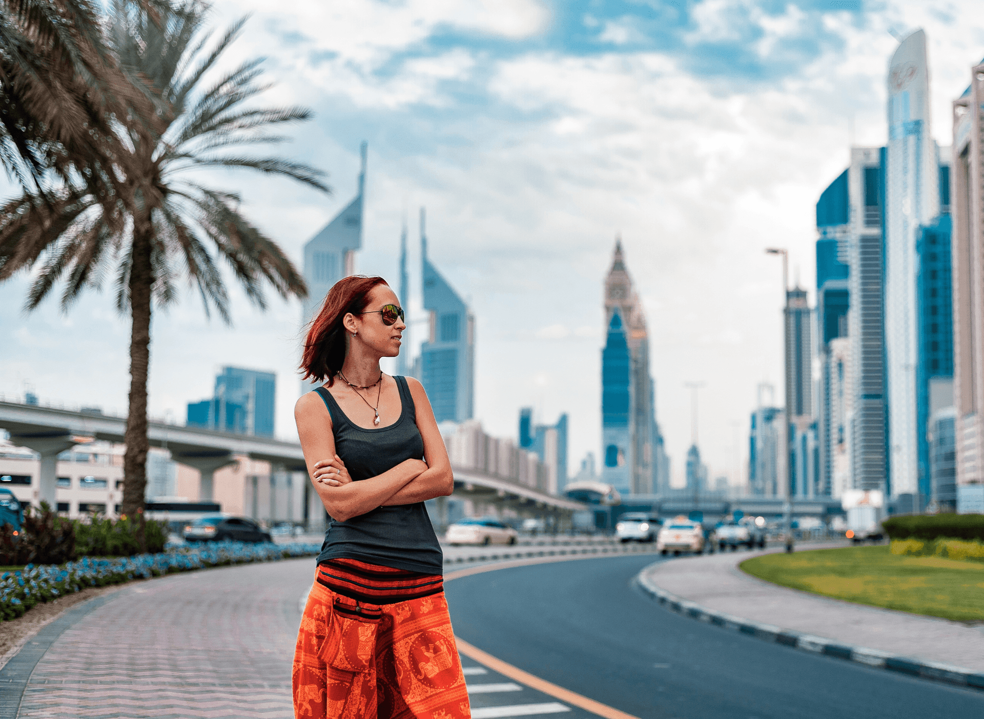 Woman with sunglasses stands on a city street, with skyscrapers and palm trees in the background.